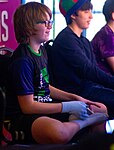 Gibson facing right in a chair playing Tetris, with the controller rested on his right foot balanced against his left knee