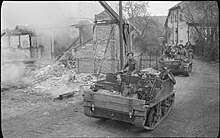 Photo noir et blanc de deux petits véhicules à chenille transportant des soldats au milieu de maisons en ruine.
