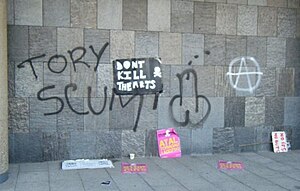 Anti-Conservative graffiti -- the words "Tory scum" next to an image of a penis -- on a wall of 30 Millbank, created during the protest. A spray-painted anarchist symbol can also be seen, along with a poster displaying a skull and crossbones with the words "Don't kill the arts", and discarded placards, one reading "No ifs, no buts" (part of a slogan ending "no Tory cuts"). Tory Scum.jpg