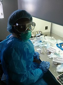 Une jeune femme à une table de travail couverte de médicaments, portant un ensemble complet de protections y compris gants, masque, lunettes.