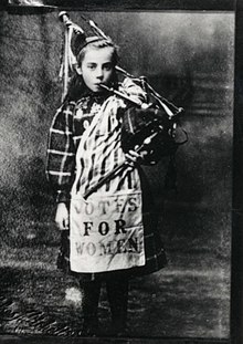 Photograph of Elizabeth Watson at the age of 9, holding a bagpipes and wearing a scarf that says "Votes for women"