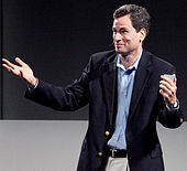 White man wearing a suit standing with his hands held far apart, looking left of image.