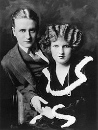A black and white portrait of F. Scott Fitzgerald and Zelda Sayre. Both are partially reclined with Zelda leaning against Fitzgerald. His right hand is clasping her left hand.