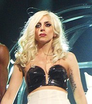 Upper torso of a young blond woman. Her hair falls in waves up to her shoulders. She wears a black shiny bustier and a white wrapping is visible underneath it. On her left hand, just below her underarm, a tattoo is visible.