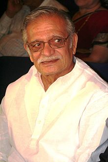 An old brown person with short hair and wearing rectangle glasses, and white shirt. He is smiling away from the camera.