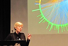 A woman with short blonde hair stands in front of a screen displaying a colorful circular network diagram