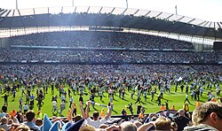 A jubilant scene at a full to capacity football stadium. Hundreds of supporters bedecked in blue are running onto the pitch in celebration