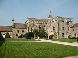 An irregular stone building of one, two and three storeys, the taller parts buttressed and embattled. Mullioned windows. One turret with a small spire. In front of the buildings a lawn.