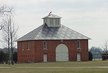 The image shows a large brick barn with a large arched front door and arched windows around two sides of the building.