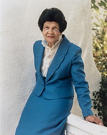 A woman with dark hair wearing a blue suit, leaning on a railing.