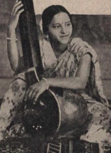 A young South Asian woman wearing a light-colored sari, seated, arms around a large stringed instrument