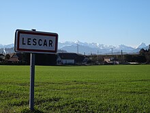 Photographie en couleurs d'un panneau avec l'inscription " Lescar", en fond des montagnes enneigées.