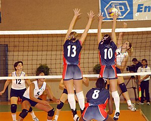 Volley-ball : l'équipe en bleu tente un contre face au smash d'une joueuse en blanc ; de part et d'autre, les autres joueuses sont en soutien.