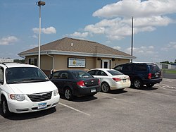 A small brown single-story building sits behind a row of cars on a sunny day.