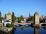 A pair of rectangular medieval stone towers guard both banks of a river and the stone bridge that stretches between them.