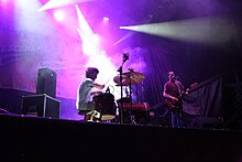 a guitarist and drummer perform on a stage under spotlights with dry ice