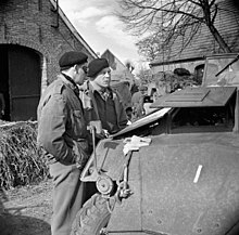 Photo noir et blanc montrant deux hommes en uniforme en train de discuter entre des maisons, près d'un véhicule blindé.
