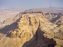 Ruins on the flat top of a sand colored mountain, surrounded by desert. Other mountains are visible in the background.