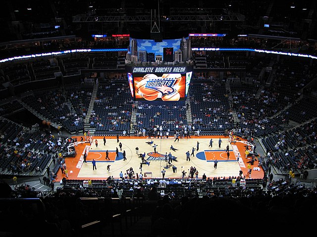 An arena packed with spectators. A basketball court is in the center, and a scoreboard is directly above it.