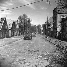 Photo en noir et blanc montrant une rue dans une ville, bordée par des maisons, avec des chars sur la route. Les bâtiments sont en bon état malgré quelques impacts..