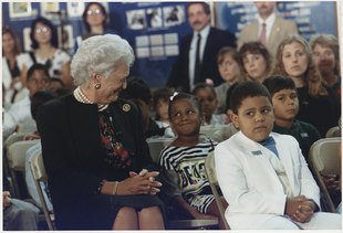 Барбара Буш и молодая девушка, сидящие рядом друг с другом в комнате, полной людей, улыбаются друг другу.