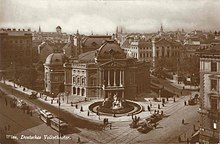 Vogelperspektive auf das Deutsche Volkstheater in Wien mit seinem klasssizistischem Säuleneingang. Auf dem Vorplatz befindet sich ein Springbrunnen mit einer Statue. Auf den sich Straßen vor dem Platz sind alte Straßenbahnen und Pferdekutschen zu erkennen.