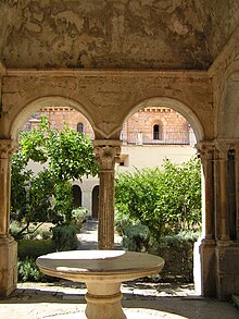 Photographie couleur d'une vasque en pierre située sous les arcades d'un cloître donnant sur un jardin.