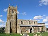 A stone church seen from the south. On the left is a battlemented tower with a semicircular stair turret, in the middle is the nave with a clerestory and a porch, and to the right at a lower level is the chancel