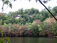 Утес с небольшим укрытием на вершине, вид через озеро. Листья на деревьях, растущих на склонах, зеленые, розовые, пурпурные и красные.