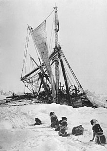 Side of a wooden steamship held in solid ice, leaning heavily to the left with a lfeboat swinging in its davits. One man visible on the ice, another aboard the ship, looking down.