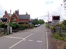 a normal country road passes straight up the middle of the picture, and a two-track railway line crosses it in the foreground. Modern red-and-white barriers protect the track, and are currently up. On the left, just beyond the line, 19th century red brick buildings stand next to the track. This was once the railway station but is now a private house. The line and the road are fringed with hedgerows, those along the road on the left being particularly tall. A field of wheat can just be glimpsed on the right, behind the barriers, lights, and hedgerows.