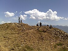 Photo en couleur montrant des hommes en haut d'une colline pierreuse, un drapeau vert et blanc est tenu par un d'eux, le ciel est bleu avec des nuages