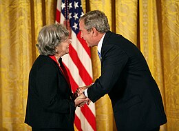 Ruth R. Wisse steht links von George W. Bush. Beide sind dunkel gekleidet. Sie schauen sich an, lächeln und drücken sich die Hände. Im Hintergrund steht eine US-amerikanische Flagge vor einem goldfarbigen Vorhang.