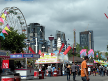 Crowds of people wander around booths selling carnival food. A merry-go-round is in the foreground to the left, and several skyscrapers stand in the background.
