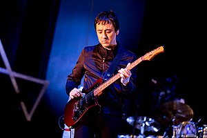 Jeff Schroeder wearing a dark zippered jacket, playing electric guitar onstage, with a blurry drumkit visible in the background