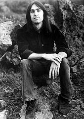 A long-haired man wearing a dark shirt and jeans, sitting on a rock in the countryside