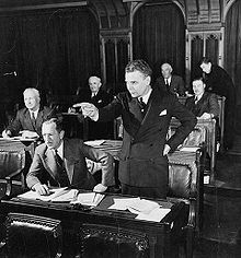 Diefenbaker, standing in a legislative chamber, dramatically points in front of him. His hair is greying, and he appears much the way he will as Prime Minister.