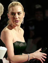 A young woman in formal evening attire of a  strapless black gown, with diamond earrings and bracelet, looks over her  shoulder. Her hair is pulled back from her face, and her hand, with  dark nail varnish clasps a purse.