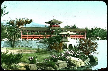 Long red wooden covered bridge with ornamental roof; woods on either side of bridge, woods and mountain behind; water and landscaped point of land with flowers in foreground