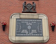 A stone tablet, inscribed with the image of a football pitch and several names. It is surrounded by a stone border in the shape of a football stadium. Above the tablet is a wooden carving of two men holding a large wreath.