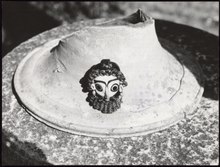 Photographie en noir et blanc d'une amulette de pâte de verre posée sur une céramique partiellement conservée