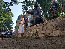 Zdjęcie pomnika Misia Wojtka w Sopocie. Rzeźba przedstawia niedźwiedzia w pozycji siedzącej, ubranego w wojskowy mundur i machającego prawą łapą. Zdjęcie wykonane podczas odsłonięcia pomnika. Po bokach stoją wyprostowani żołnierze z karabinami. Przed murkiem, na którym stoi pomnik, przemawia prelegentka, a na krzesłach obok siedzi publiczność, wśród której widać weterana wojennego