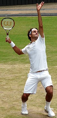 A dark-haired man is in the serving motion, which he is in all white clothing, and he has a redish-black tennis racket in his right hand
