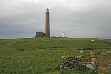 A tall, conical grey tower with outbuildings sits in a green field. A second small, white tower is in the background.
