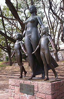 Photographie en couleur d'une statue en bronze sous des arbres, posée sur un socle en pierre et représentant une femme entourée de deux enfants.