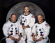 Photographie couleur de l'équipage d'Apollo 11 en combinaison Apollo avec la Lune en fond.