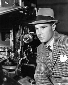 A man in a suit and hat next to a camera.