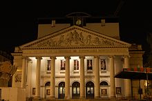 Photographie de nuit de la façade éclairée du théâtre : le péristyle aux huit colonnes surmontées du fronton sculpté précédant l'entrée du grand hall.