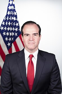 Hispanic man in a black suit and red tie with an American flag in the background.