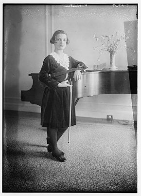 A young woman standing beside a piano, holding a violin and bow, looking at the camera; she has dark glossy hair cropped to jaw length, and is wearing a dark dress with a white lace collar.